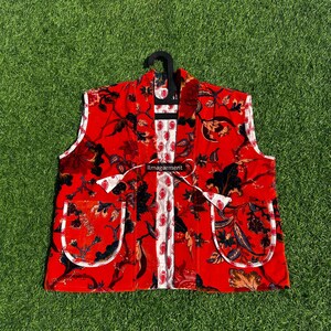 May include: A red vest with a floral pattern, white trim, and two front pockets. The vest is hanging on a black hanger against a green grass background. The text "ilmagarment" is visible.