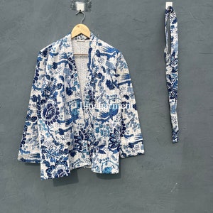 Op de afbeelding: Een wit en blauw gepatroneerd jasje in kimono-stijl met een bijpassende band. Het jasje heeft een bloemen- en vogelmotief.