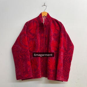Könnte beinhalten: Rote Samtjacke mit aufwendiger Blumenstickerei. Die Jacke hat einen hohen Kragen und eine Knopfleiste. Die Stickerei ist in einem dunkleren Rotton gehalten und bildet ein kontrastreiches Design. Der Text "llmagarment" ist sichtbar.
