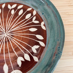 May include: A handcrafted ceramic plate with a teal rim and a brown centre. The centre features a white floral design. The plate is round and glazed, with a rustic, artisanal aesthetic.