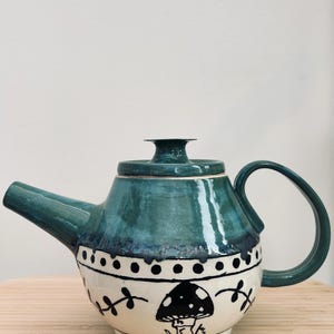 May include: A teal ceramic teapot with a mushroom design. The teapot has a rounded body with a white band decorated with black dots and a mushroom illustration. The handle and spout are teal, and the lid is teal with a small knob.