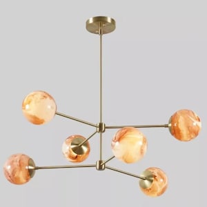 May include: A modern chandelier with a brass finish and six globe-shaped shades. The shades have a marbled, orange and cream color pattern. The fixture is suspended from a ceiling mount.