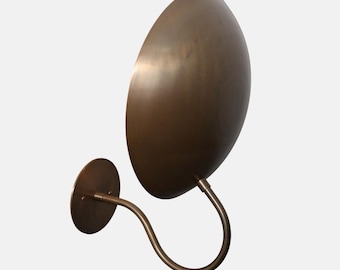 May include: A bronze-colored wall sconce with a large, oval shade. The shade is attached to a curved arm and a circular base. The fixture has a minimalist design and a warm, metallic finish, suitable for interior lighting.