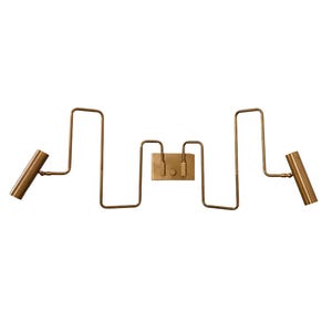 May include: A brass-toned wall-mounted light fixture with a unique, geometric design. The fixture features two cylindrical spotlights on either end, connected by a series of angular, zig-zagging tubes to a central rectangular plate with two small switches.