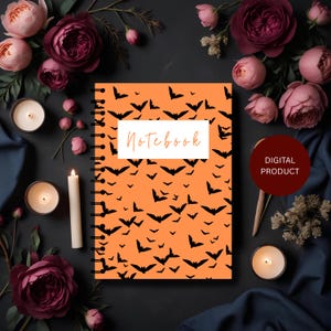 Op de afbeelding: Oranje notitieboek met een zwart vleermuismotief en het woord "Notebook" in een witte rechthoek. Het notitieboek heeft een spiraalbinding. De afbeelding is omgeven door bloemen, kaarsen en een donkerblauwe stof.