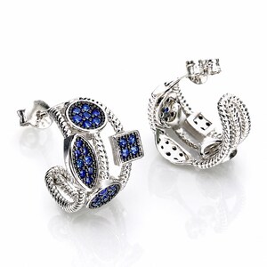 May include: A pair of silver hoop earrings featuring a twisted rope design. Each earring is adorned with multiple geometric settings, including circular, oval, and square shapes, all inlaid with small, vibrant blue stones and black accents.