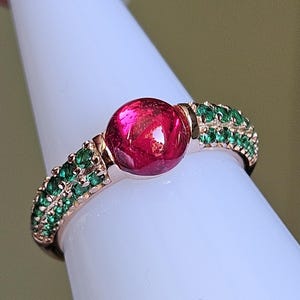 Peut inclure: Une bague en or rose ornée d'une grosse pierre rouge ronde au centre. La bande est ornée de rangées de petites pierres vertes. La bague est présentée sur un objet blanc en forme de cône.
