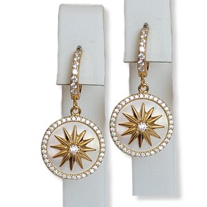 Puede incluir: Aretes de aro dorados con un disco blanco de nácar colgante que presenta un diseño de estrella dorada con detalles de cristal transparente.