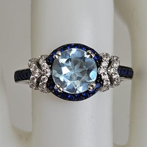 Puede incluir: Un anillo de plata con una gran piedra preciosa azul claro rodeada por un halo de piedras azules más pequeñas y una banda de piedras blancas más pequeñas.