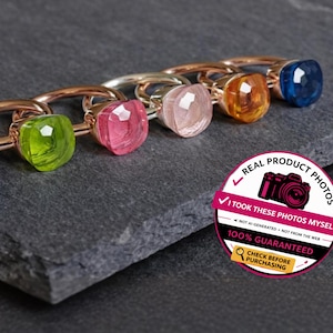 Può includere: Una collezione di anelli con pietre preziose colorate, tra cui blu, verde, rosa e arancione, esposti su una superficie grigio scuro. Gli anelli hanno montature dorate o argentate e pietre quadrate. Un adesivo con testo è visibile.