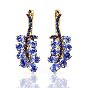 Op de afbeelding: Een paar gouden oorbellen met een gebogen steeltje bezet met kleine donkerblauwe steentjes. Elke oorbel is versierd met meerdere ovaalvormige lichtpaarse edelstenen die in een tros langs de zijkant van het gouden steeltje zijn geplaatst.