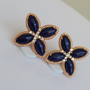 May include: A pair of gold earrings with four-leaf clover designs. Each clover has four dark blue oval-shaped stones and a cluster of small clear stones in the center.