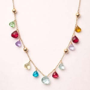Könnte beinhalten: Eine zarte goldene Halskette mit tropfenförmigen Edelsteinen in verschiedenen Farben, darunter Grün, Rot, Blau und Gelb. Die Halskette hat kleine goldene Perlen entlang der Kette, die ein farbenfrohes und elegantes Design schaffen.