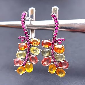 May include: A pair of silver earrings featuring a curved stem paved with small pink stones. Attached to the stems are clusters of oval-cut gemstones in shades of yellow, orange, and light green, creating a vibrant, multi-coloured fruit-like design.