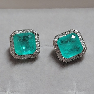 Puede incluir: Un par de pendientes de plata con una piedra preciosa cuadrada verde esmeralda rodeada por un halo de piedras preciosas más pequeñas y transparentes.