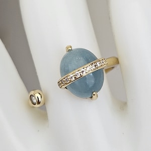 Op de afbeelding: Een gouden ring met een lichtblauwe ovale edelsteen gezet in een gouden band met een rij heldere, glinsterende stenen. De ring heeft een delicaat open bandontwerp.