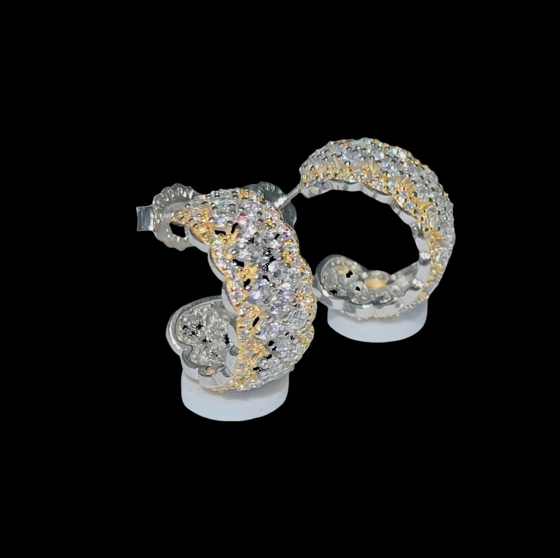May include: A pair of silver-toned hoop earrings with a gold-colored inner design. The earrings are encrusted with numerous small, clear gemstones, creating a sparkling effect. The earrings are displayed on a white surface against a black background.