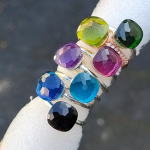 Könnte beinhalten: Eine Sammlung von Edelsteinringen in verschiedenen Farben, darunter Blau, Lila, Grün und Rosa. Jeder Ring hat einen quadratisch geschliffenen Edelstein auf einem Silberband. Die Ringe sind gestapelt angeordnet und zeigen ihre lebendigen Farbtöne.