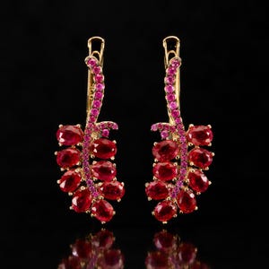 May include: A pair of gold dangle earrings featuring a vertical stem encrusted with small pink stones, with multiple oval-cut red gemstones arranged in a cluster resembling a bunch of grapes hanging from the bottom of each earring.