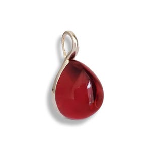 Puede incluir: Colgante en forma de lágrima con una gema roja vibrante. La gema está engastada en un marco de metal plateado y tiene un lazo en la parte superior para una cadena. El colgante se muestra sobre un fondo blanco.