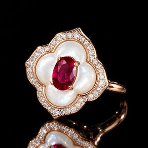 Può includere: Un anello in oro rosa con un rubino rosso centrale a taglio ovale, circondato da un inserto in madreperla bianca a quattro lobi. Il bordo esterno della montatura è decorato con piccoli diamanti bianchi rotondi su fascia in oro rosa.