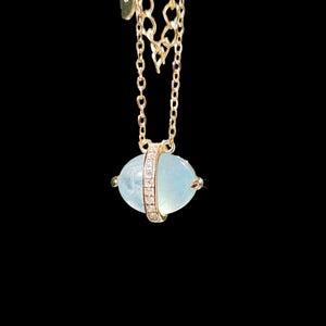 May include: A gold-colored necklace with a light blue, oval-shaped pendant. The pendant is encircled by a band of small, clear stones. The necklace chain is delicate and also gold-colored, set against a black background.