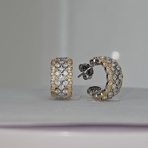 May include: A pair of hoop earrings with a gold and silver design. The earrings are encrusted with small, sparkling stones. The gold sections are on the edges of the earrings. The earrings are on a white surface.