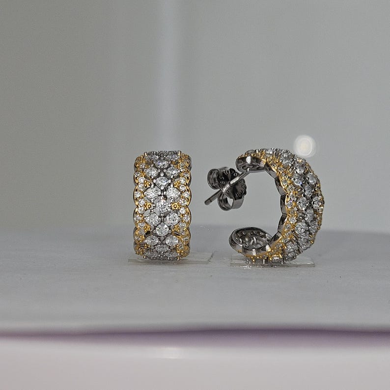 May include: A pair of hoop earrings with a gold and silver design. The earrings are encrusted with small, sparkling stones. The gold sections are on the edges of the earrings. The earrings are on a white surface.
