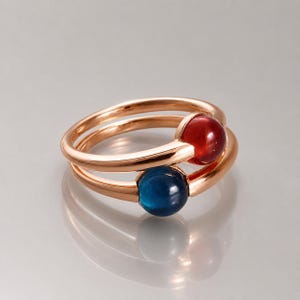 Peut inclure: Une bague en or rose avec deux anneaux, chacun tenant une pierre précieuse ronde. Une pierre est rouge foncé, l'autre bleu profond. Le design de la bague est moderne et élégant, adapté au quotidien ou aux occasions spéciales.