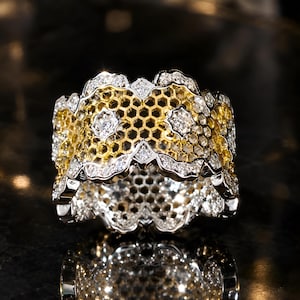 May include: A wide band ring featuring a yellow gold honeycomb pattern layered beneath a silver-toned metal frame. The design is accented with numerous small, clear, sparkling stones set along the scalloped edges and within floral-shaped motifs.