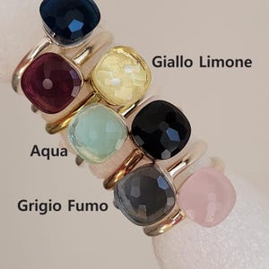 Puede incluir: Una colecci&oacute;n de anillos de piedras preciosas en varios colores, incluyendo azul marino, burdeos, amarillo, aguamarina, negro, gris y rosa. Cada anillo presenta una piedra de corte cuadrado engastada en una banda dorada. El texto "Giallo Limone", "Aqua" y "Grigio Fumo" es visible.