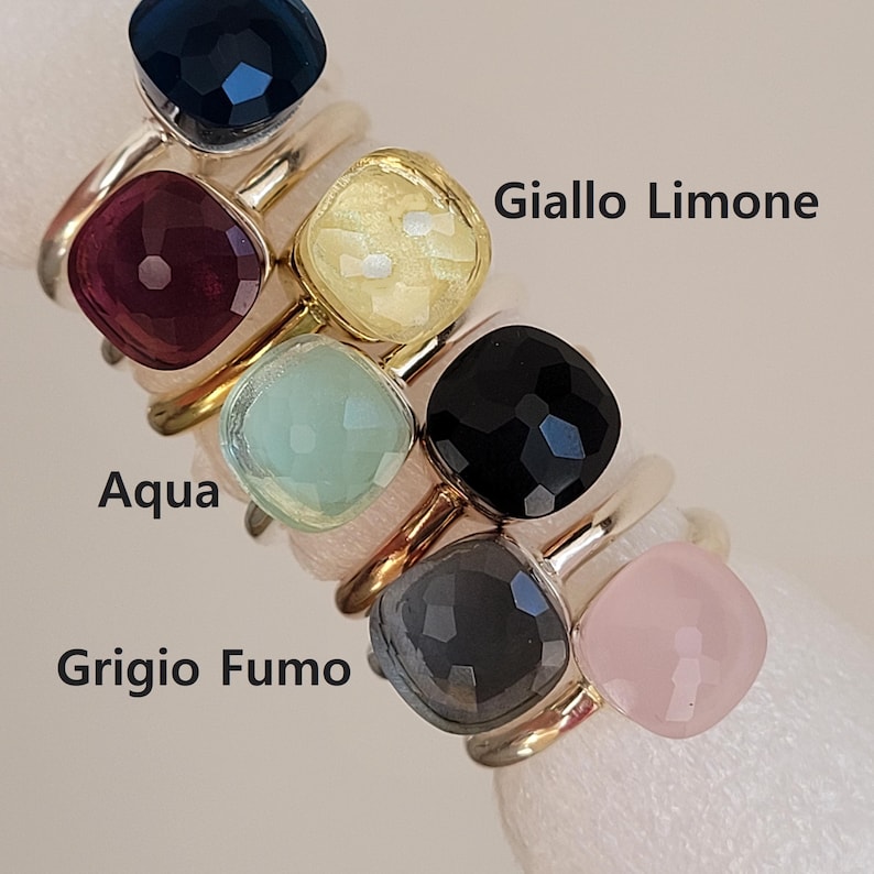 Puede incluir: Una colecci&oacute;n de anillos de piedras preciosas en varios colores, incluyendo azul marino, burdeos, amarillo, aguamarina, negro, gris y rosa. Cada anillo presenta una piedra de corte cuadrado engastada en una banda dorada. El texto "Giallo Limone", "Aqua" y "Grigio Fumo" es visible.