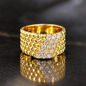 May include: A wide gold band ring featuring a textured surface of small, polished gold spheres arranged in rows. A diagonal section of the ring is set with numerous clear, round-cut white stones that contrast with the metallic gold finish.