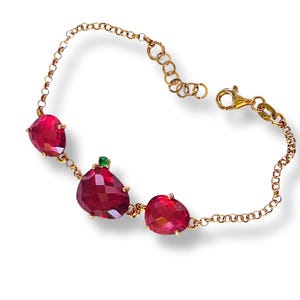 May include: A gold-toned bracelet featuring three large, faceted, red gemstones and a small green gemstone. The bracelet has a delicate chain design and a lobster clasp closure. The gemstones are set in gold-colored prongs.