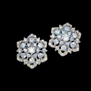 Puede incluir: Un par de pendientes de botón brillantes. Cada pendiente presenta un diseño floral con numerosas piedras preciosas redondas y transparentes engastadas en metal dorado. Fondo negro.