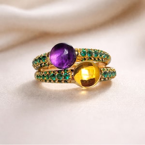 Puede incluir: Dos anillos dorados, uno encima del otro, cada uno con una gema grande y redonda. El anillo superior tiene una gema púrpura, el inferior una amarilla. Ambos anillos están adornados con pequeñas gemas verdes.