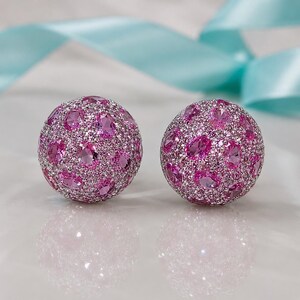 Könnte beinhalten: Ein Paar kugelförmige Ohrstecker mit einer dichten Pavé-Fassung aus kleinen, klaren weißen Kristallen, durchsetzt mit größeren, oval geschliffenen, leuchtend rosa Edelsteinen. Die Oberfläche jeder Kugel ist vollständig mit diesen Steinen besetzt.