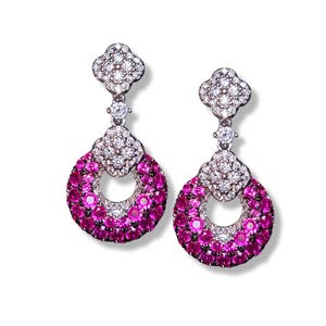 Puede incluir: Un par de pendientes colgantes con una parte superior en forma de flor y una gota circular. Los pendientes están adornados con pequeñas piedras preciosas redondas blancas y rosas. Las piedras están engastadas en un metal plateado.
