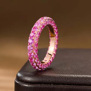 Könnte beinhalten: Ein Ewigkeitsring aus Roségold mit zwei parallelen Reihen aus runden, leuchtend pinken Edelsteinen, die rund um den gesamten Ring gefasst sind. Der Metallring hat eine polierte Oberfläche, die die hellen pinken Steine vor dem warmen Goldton hervorhebt.