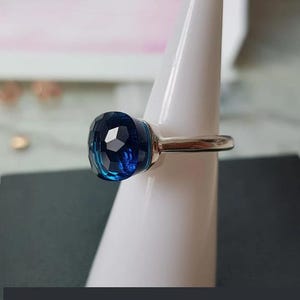 Puede incluir: Un anillo de plata con una gran gema facetada de color azul oscuro. La gema tiene forma redondeada de coj&iacute;n y refleja la luz, creando un efecto brillante. El anillo se exhibe sobre un soporte blanco.