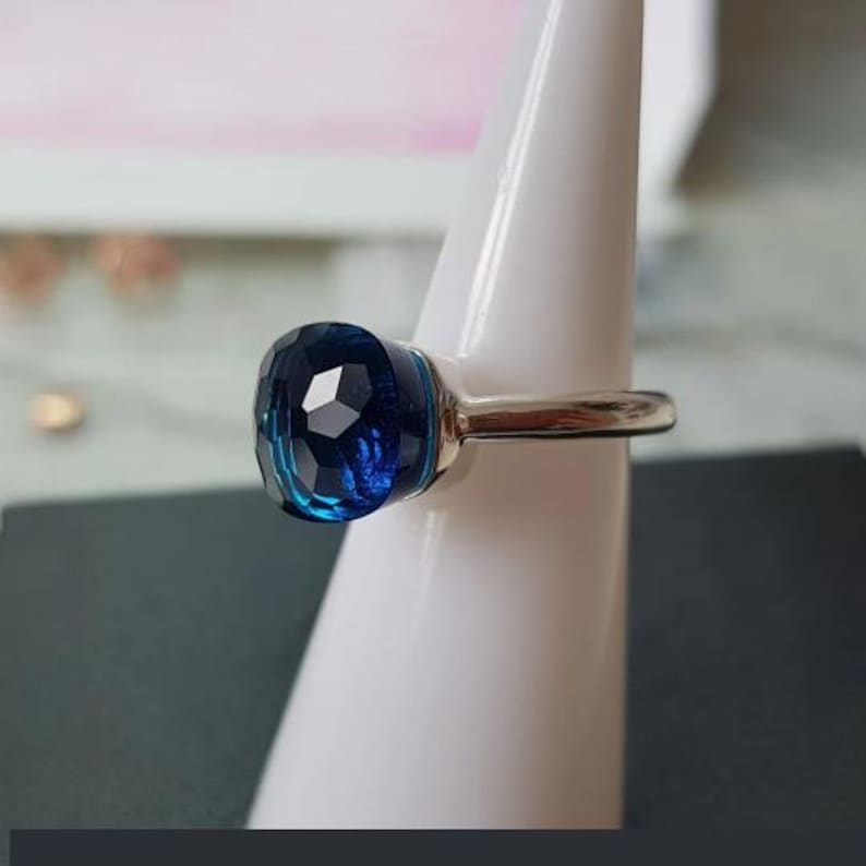 Puede incluir: Un anillo de plata con una gran gema facetada de color azul oscuro. La gema tiene forma redondeada de coj&iacute;n y refleja la luz, creando un efecto brillante. El anillo se exhibe sobre un soporte blanco.
