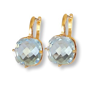 Puede incluir: Un par de pendientes de oro con cierre de palanca que presentan grandes piedras preciosas de color azul claro, talladas en forma cuadrada y facetadas. Cada piedra está engastada en un marco de oro pulido con cuatro garras.