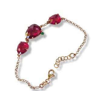May include: A gold chain bracelet with three red gemstone charms. The bracelet has a lobster clasp closure.