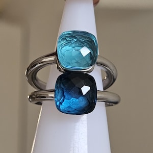 Könnte beinhalten: Zwei Ringe mit quadratischen, facettierten blauen Edelsteinen. Der obere Ring hat einen hellblauen Stein, der untere einen dunkleren blauen Stein. Beide Ringe haben silberne Bänder und werden auf einem weißen Ständer präsentiert.