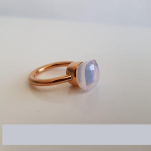 Puede incluir: Un anillo de oro rosa con una gema opalescente grande, de corte cuadrado. La gema tiene un aspecto suave y lechoso con sutiles tonos azules y rosas. La banda del anillo es de dise&ntilde;o simple y pulido.