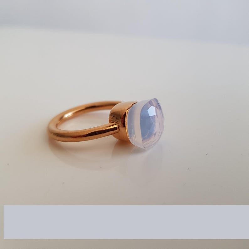 Puede incluir: Un anillo de oro rosa con una gema opalescente grande, de corte cuadrado. La gema tiene un aspecto suave y lechoso con sutiles tonos azules y rosas. La banda del anillo es de dise&ntilde;o simple y pulido.