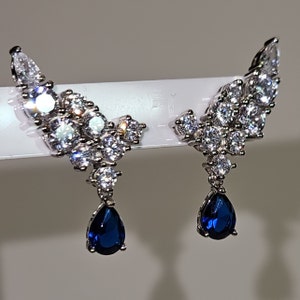 May include: A pair of silver earrings with a teardrop-shaped blue gemstone dangling from each earring. The earrings are adorned with a cluster of smaller clear gemstones.