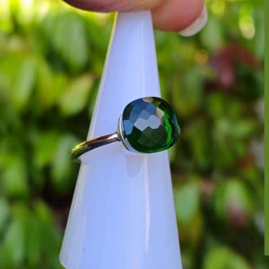Puede incluir: Un anillo de plata con una gran gema verde esmeralda facetada. El anillo se muestra sobre un objeto blanco en forma de cono, sobre un fondo verde borroso. La gema tiene un corte coj&iacute;n cuadrado.