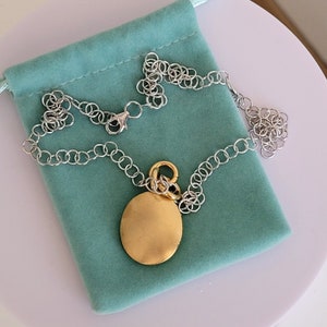 May include: A gold pendant necklace with a round, smooth, gold charm. The necklace is on a silver chain and is displayed on a turquoise velvet pouch.
