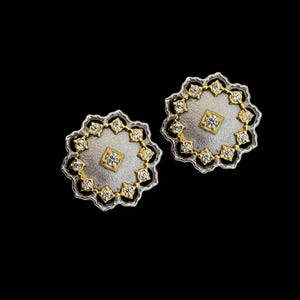 May include: Two silver and gold-tone stud earrings. Each earring features a central square-shaped gold setting with a clear stone, surrounded by a silver disc. The disc is encircled by a gold-tone border with clear stones and a scalloped edge.